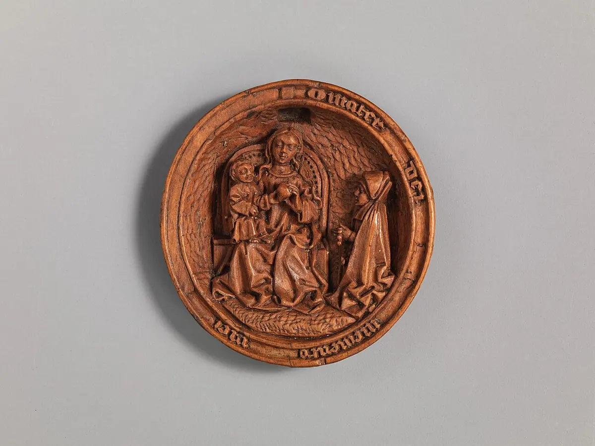 Round carved boxwood half-bead showing a tiny scene of the Virgin and Child enthroned with a kneeling figure, framed by a ring of carved lettering
