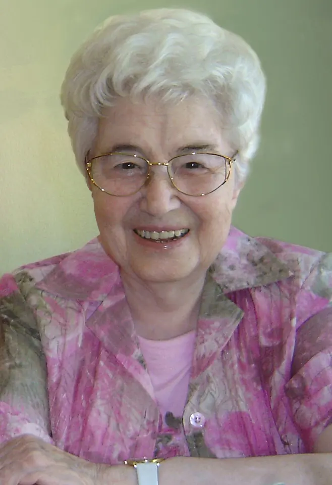 Color photograph of a smiling older woman with white hair and glasses, wearing a pink patterned blouse