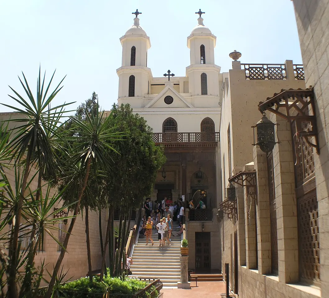 White twin bell towers above a steep stairway crowded with visitors, framed by palm trees, at the entrance of an old church