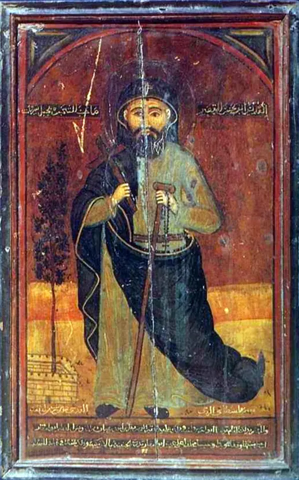 Coptic icon of a monk in a dark habit and cap with a long beard, holding a cross and a staff, before a reddish background with Arabic writing at the top