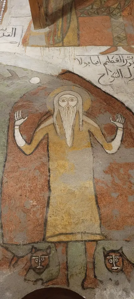 An old wall painting of a long-bearded hermit in a yellow tunic with his hands raised in prayer, two lions at his feet, and Arabic writing beside him