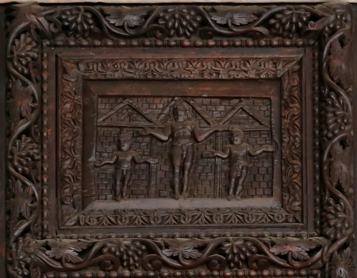 Carved wooden panel in a vine-decorated frame showing Christ with arms outstretched between two smaller figures, in front of a wall with gables