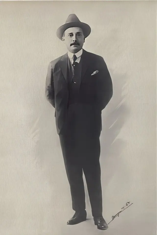Old full-length photograph of José Gregorio Hernández in a dark suit and fedora, standing with his hands behind his back