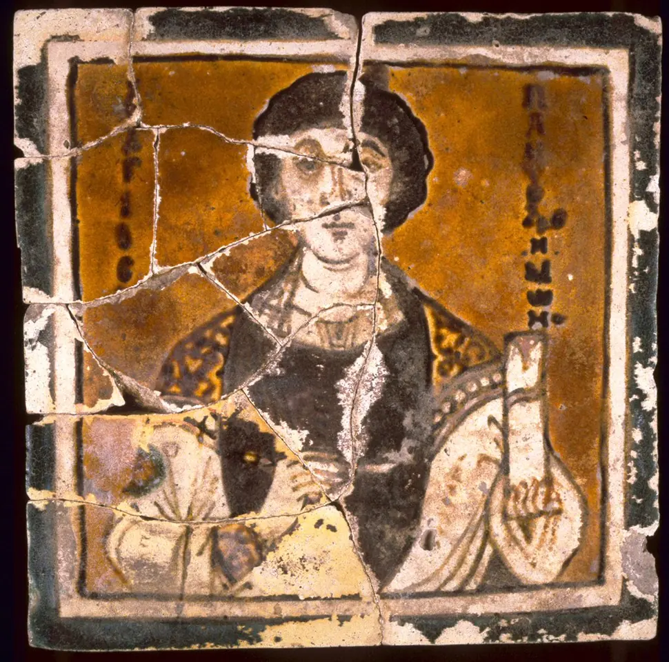 A cracked Byzantine ceramic tile with a painted bust of a young saint with dark curly hair, holding a lancet, against a yellow-brown ground with his name in Greek