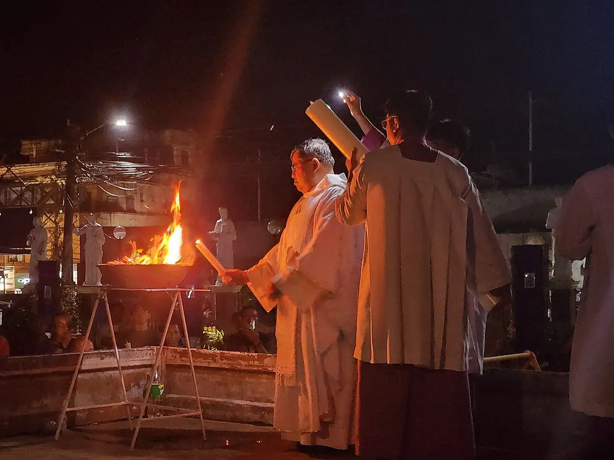 At night outside a church, a priest in white vestments stands beside a burning brazier while servers hold the tall paschal candle, ready to light it from the new fire