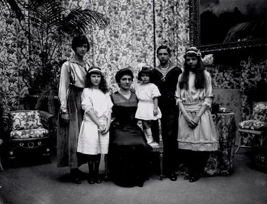 Black-and-white family photograph of a seated woman in a dark dress surrounded by her five children, three girls in white and pale dresses, a small child standing on a chair and a teenage boy in a sailor suit