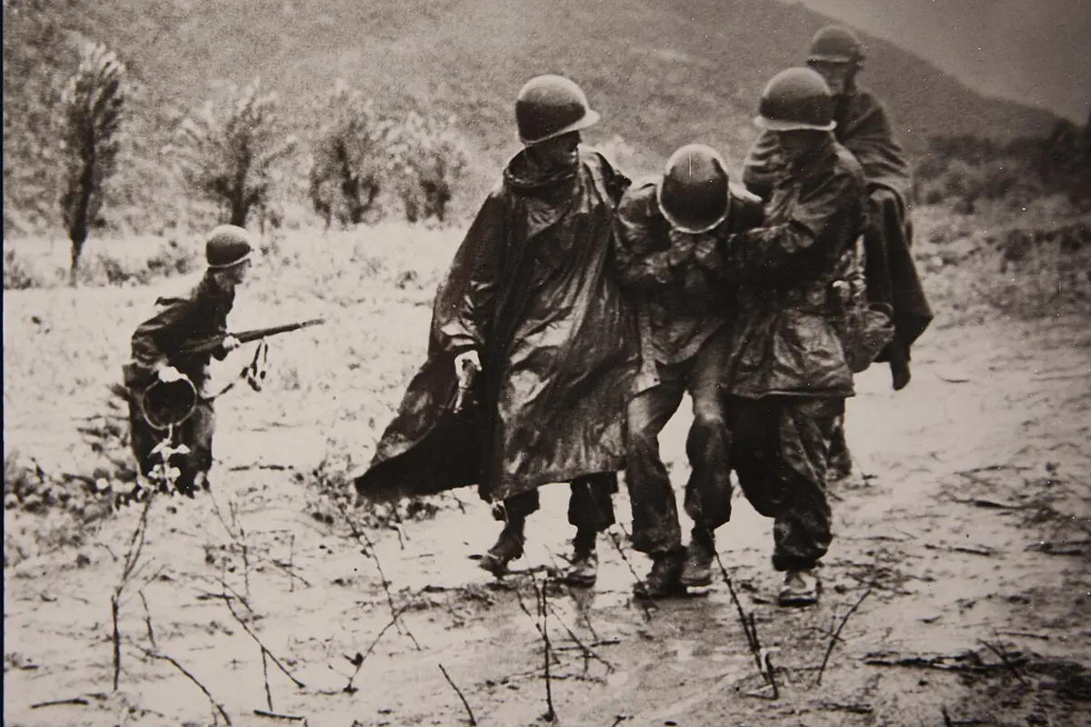 Black-and-white combat photograph of soldiers in rain ponchos and helmets half-carrying an exhausted comrade across a muddy field while another soldier crouches with a rifle