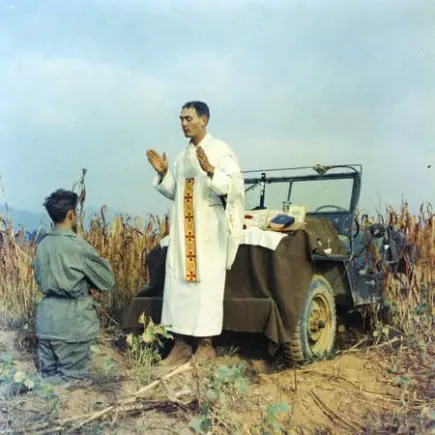 Color photograph of a priest in white vestments with a red stole praying at an altar set up on the hood of an Army jeep in a field of tall dry grass, a soldier kneeling beside him