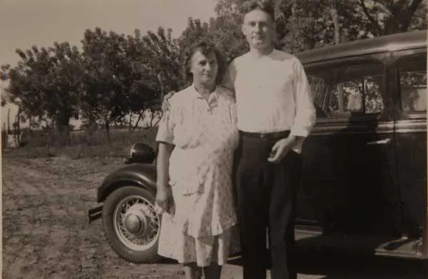 Old sepia photograph of a young man in a white shirt standing beside his mother in a print dress next to a 1930s car on a Kansas farm