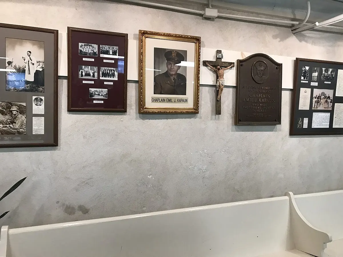 A wall in a church basement hung with framed photographs of a chaplain, a crucifix and a bronze memorial plaque