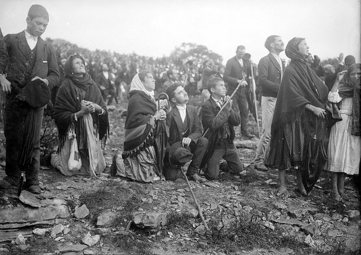 Fotografía en blanco y negro de una multitud de hombres y mujeres con sombreros y mantones, de pie y de rodillas sobre un terreno pedregoso, muchos mirando al cielo