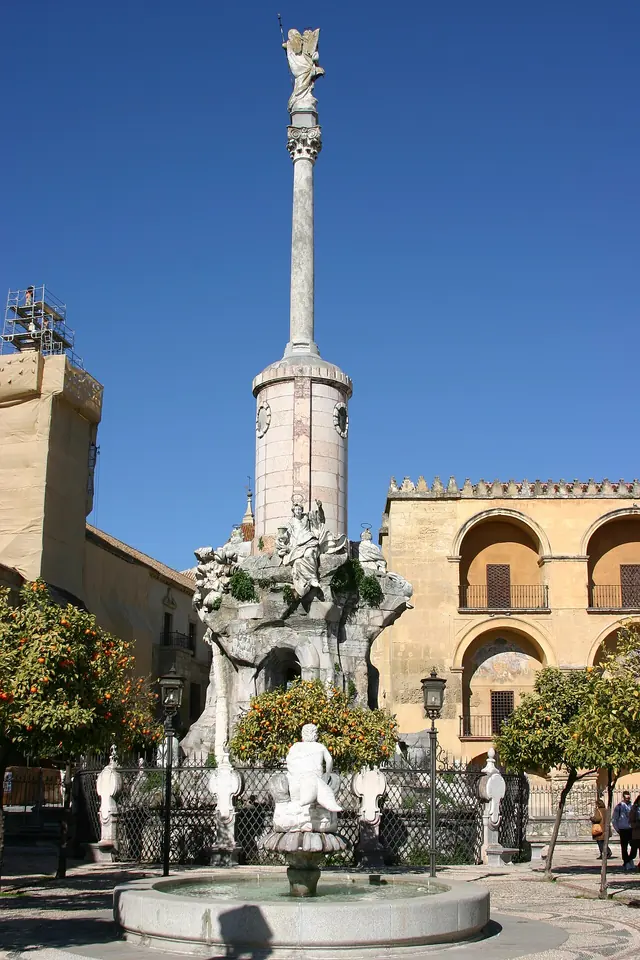 Alta columna de piedra coronada por una estatua del arcángel Rafael, sobre una base de rocas esculpidas, junto a los muros de la catedral de Córdoba bajo un cielo azul