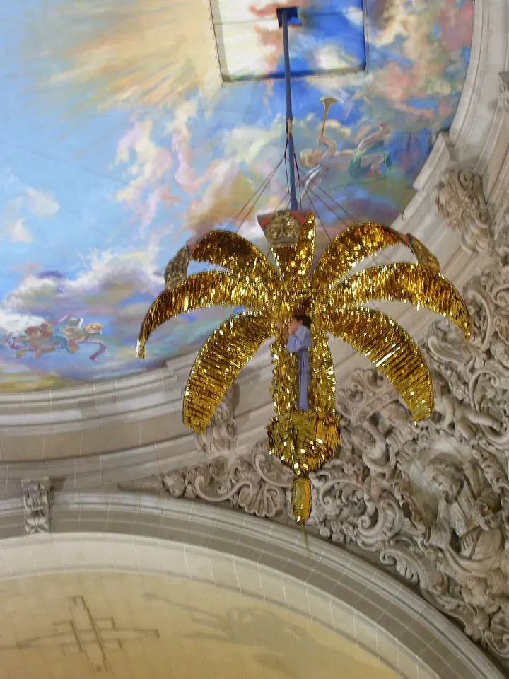 Una gran granada dorada y abierta en forma de palmera desciende desde la cúpula pintada de cielo y nubes de una basílica barroca, colgada de una cuerda