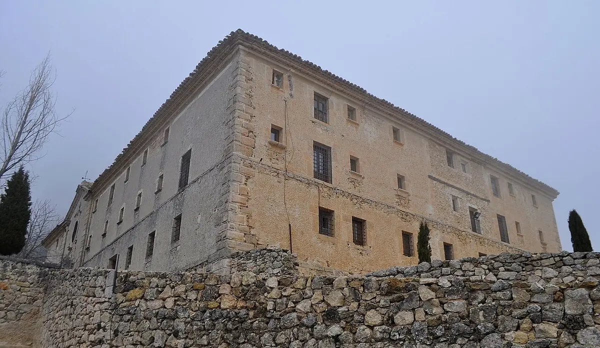 Gran edificio conventual de piedra clara, de tres pisos y ventanas pequeñas, sobre un muro de piedra seca, bajo un cielo gris de invierno