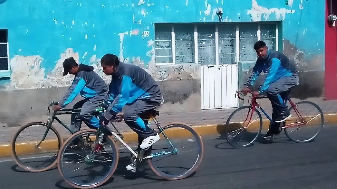 Tres ciclistas con chamarras deportivas azules pedalean por una calle de pueblo frente a una pared pintada de azul turquesa