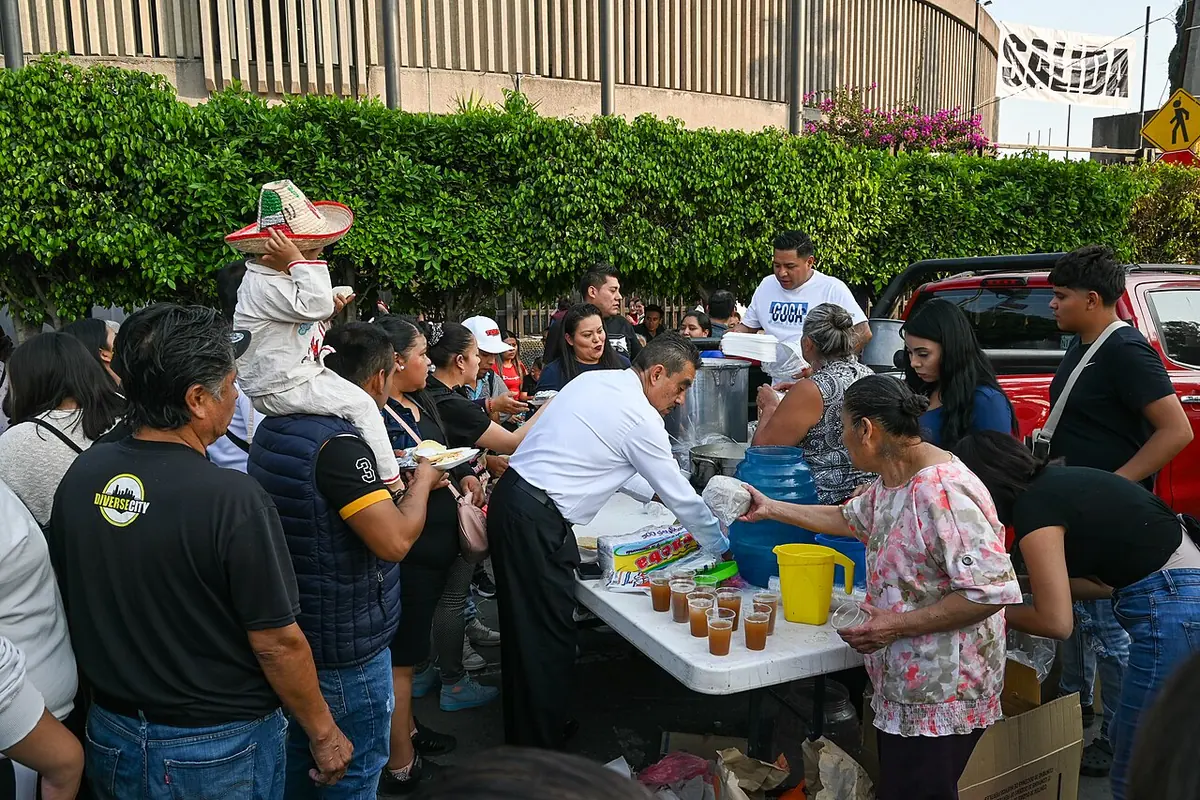 Voluntarios detrás de una mesa en la calle sirven vasos de café y comida a una fila de peregrinos, entre ellos un niño vestido de Juan Diego con sombrero sobre los hombros de su papá