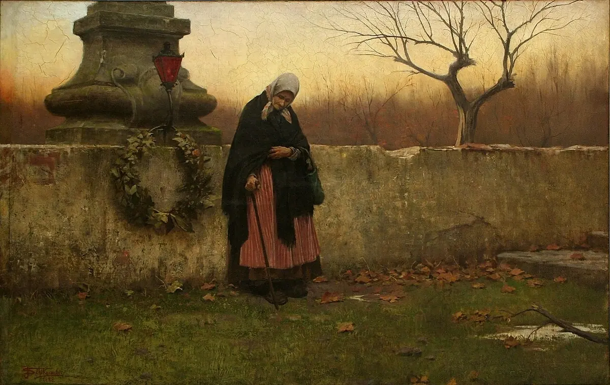 Una anciana con chal oscuro y la cabeza inclinada junto al muro de un cementerio adornado con una corona, bajo un árbol sin hojas a la luz del otoño
