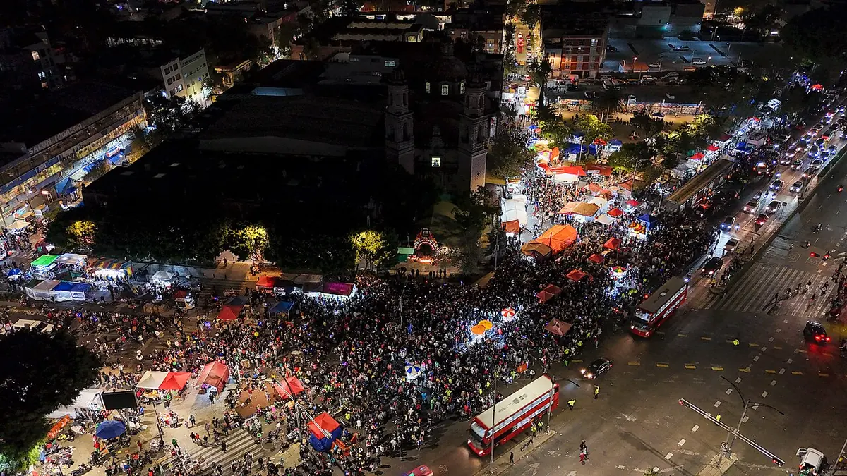Vista aérea nocturna de una enorme multitud que llena las calles alrededor del templo de San Hipólito, con toldos de puestos iluminados
