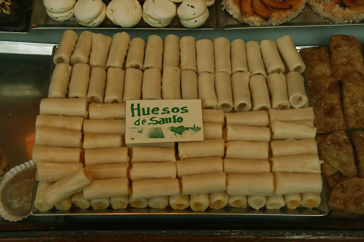 Vitrina de pastelería con filas de huesos de santo, pequeños rollos blancos de mazapán, con un cartel escrito a mano