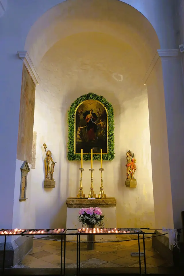 Capilla lateral blanca y abovedada con el cuadro de María Desatanudos en una corona verde sobre un altar con candeleros, flores y filas de veladoras
