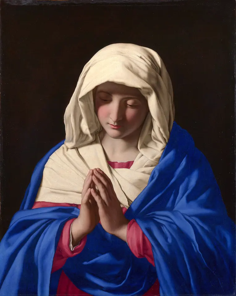 La Virgen María, de medio cuerpo, con velo blanco y manto azul intenso, inclina la cabeza y junta las manos en oración sobre un fondo oscuro