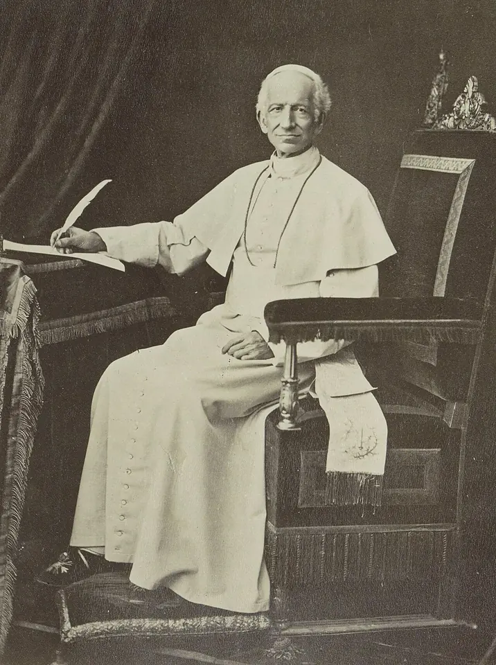 Fotografía antigua del papa León XIII con sotana blanca y solideo, sentado en un sillón junto a un escritorio con una pluma en la mano