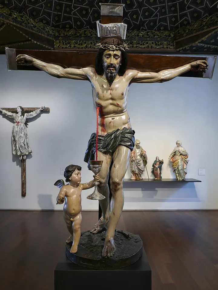 Talla barroca de madera policromada de Cristo crucificado, con la sangre que corre de sus heridas y un ángel niño a sus pies que sostiene un cáliz para recogerla