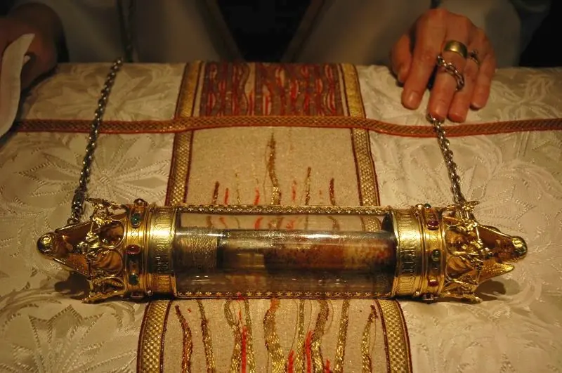 Relicario cilíndrico de cristal con remates de oro trabajado, sostenido por cadenas sobre una estola bordada, ante las manos de un sacerdote