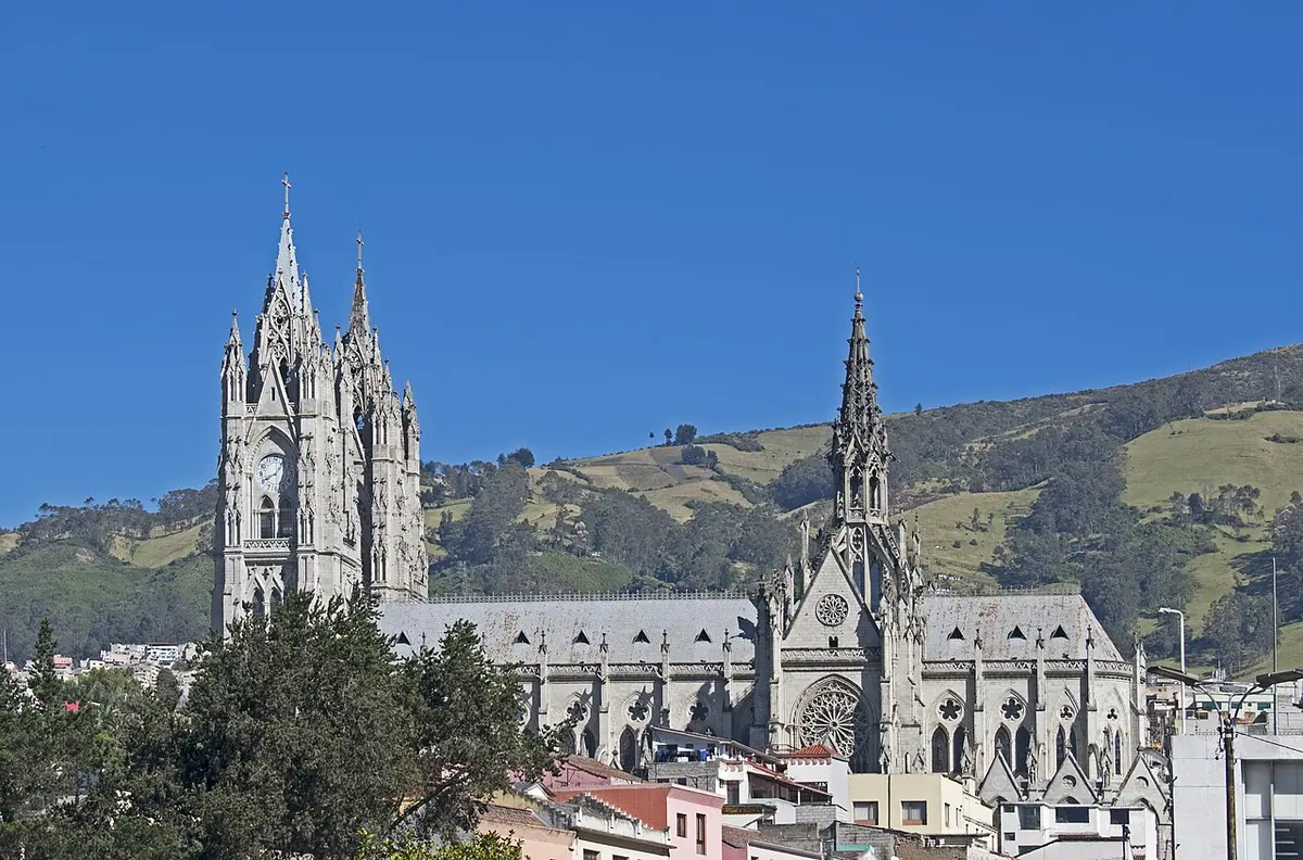 Gran basílica neogótica de piedra gris con dos altas torres caladas y una aguja, sobre los tejados de Quito y con montañas verdes al fondo