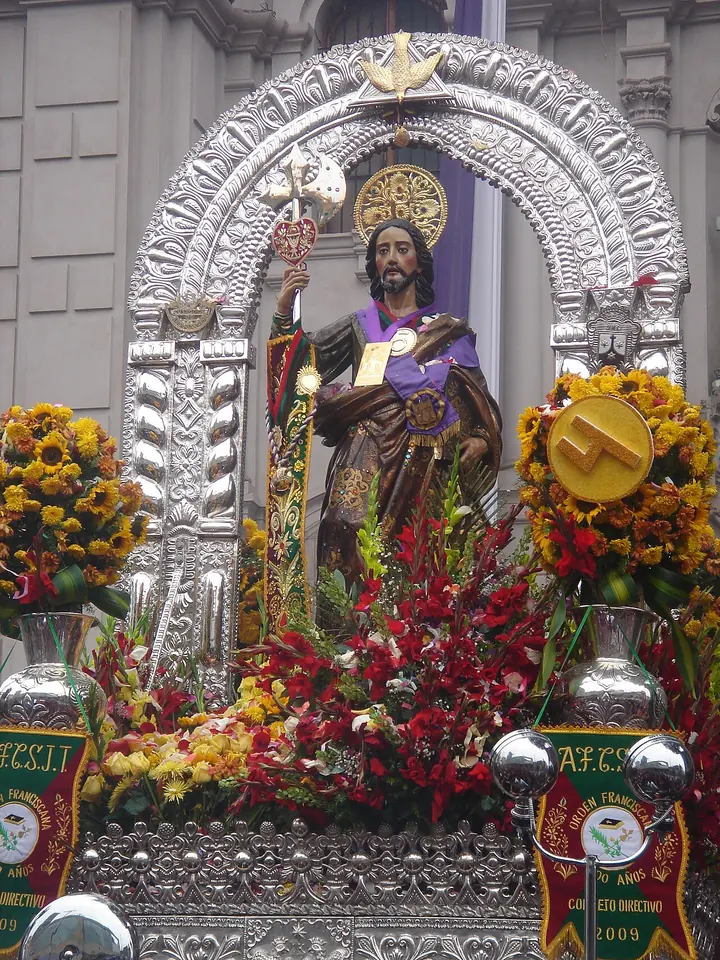 Imagen de San Judas con manto morado y blanco llevada en un anda de plata bajo un arco plateado, rodeada de flores rojas y amarillas