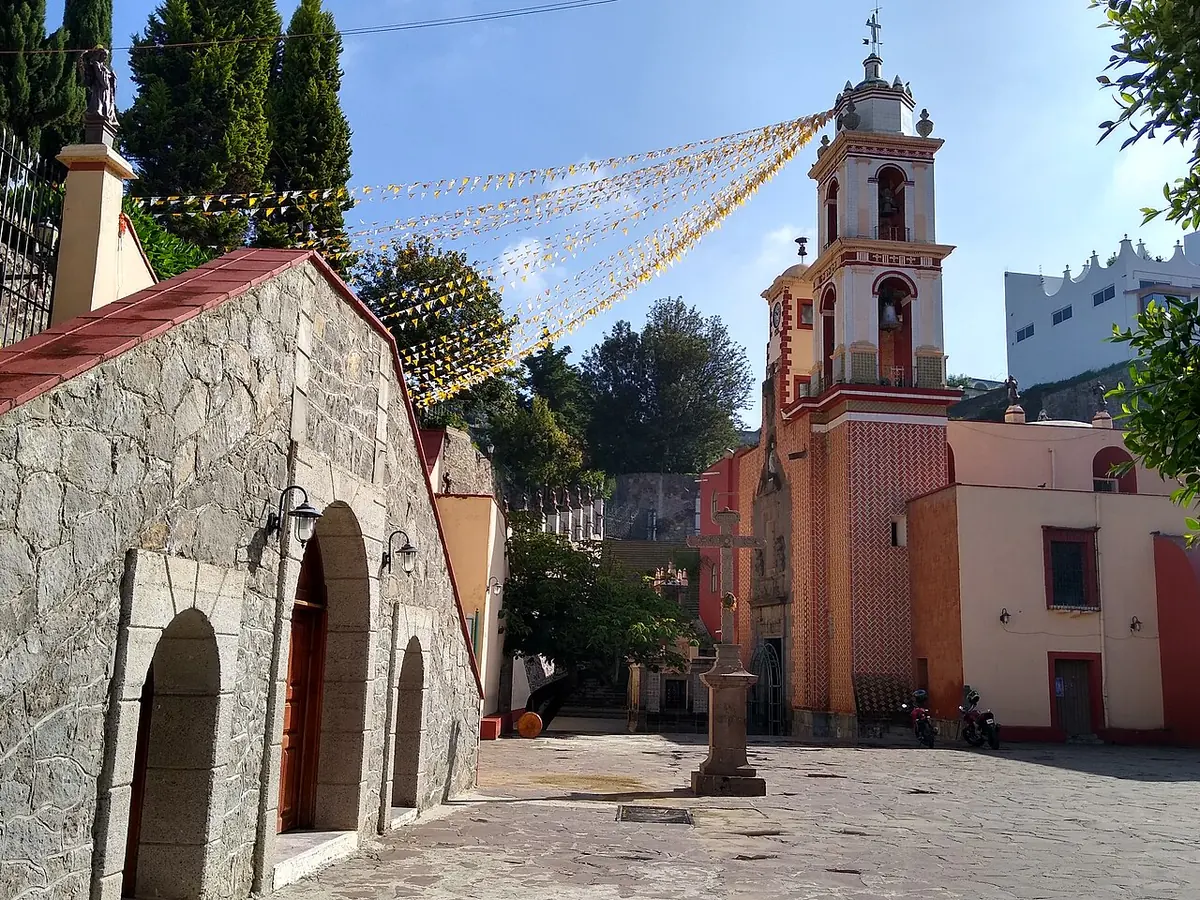 Atrio empedrado del santuario de San Miguel del Milagro, con una capilla de piedra a la izquierda, una cruz atrial y el campanario rosado adornado con banderitas de papel