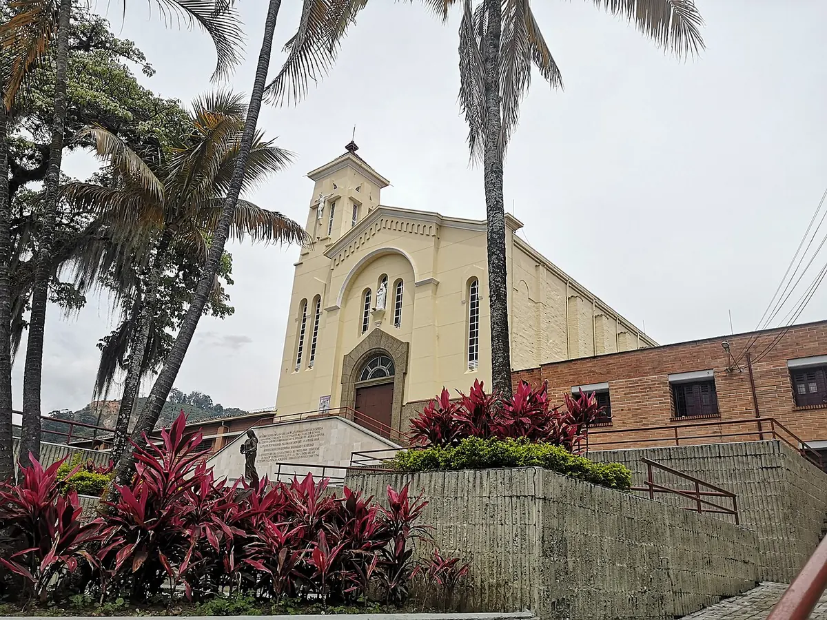 La iglesia del santuario de Santa Laura Montoya en Medellín, de fachada color crema con un campanario, sobre unas escalinatas rodeadas de palmeras y plantas de hojas rojas