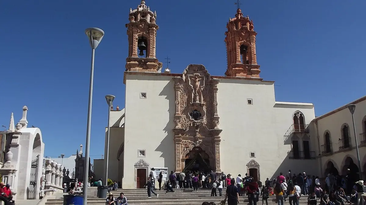Fachada barroca de cantera rosa y blanca del santuario de Plateros, con dos torres labradas, y peregrinos en la escalinata del atrio bajo un cielo azul