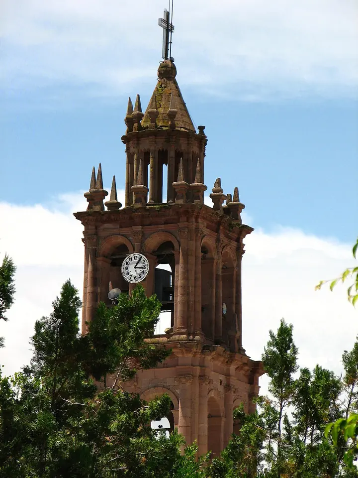 Una alta torre de cantera rosa con reloj, arcos de campanario y una cruz en lo alto, sobre árboles verdes y un cielo azul