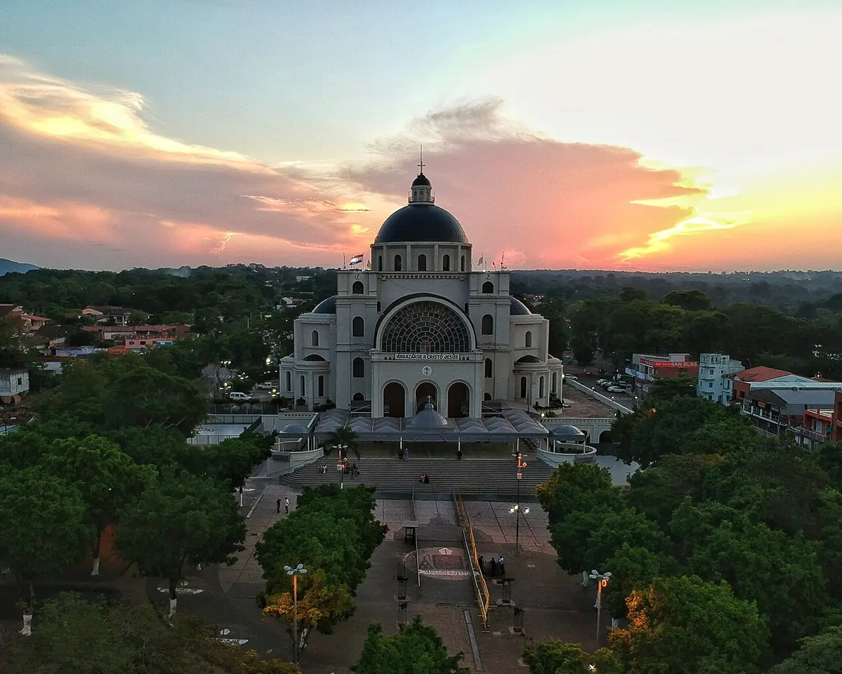 Vista aérea de la basílica de Caacupé, de color claro con una gran cúpula oscura, frente a su explanada y rodeada de árboles al atardecer