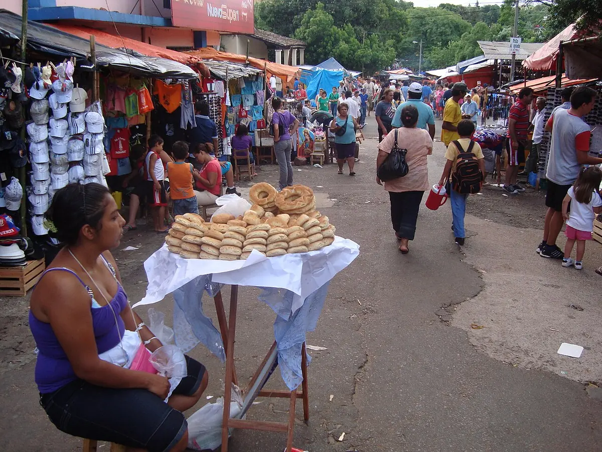 Una vendedora sentada junto a una mesita con roscas de chipa en una calle llena de puestos y peregrinos durante la fiesta de Caacupé