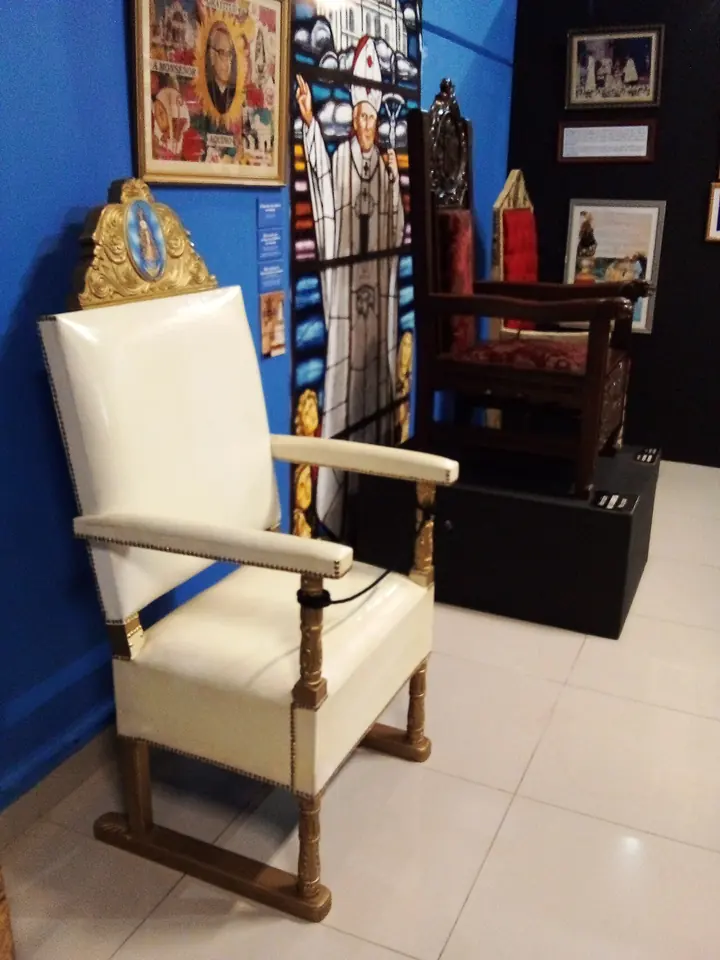 Un sillón blanco con respaldo dorado expuesto en una sala del museo de la basílica, junto a cuadros y vitrinas