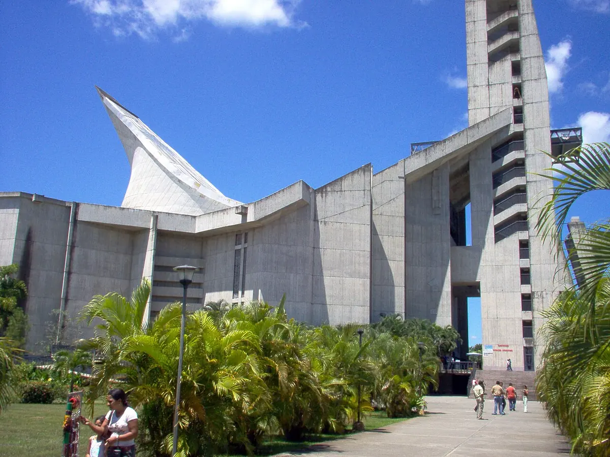 Peregrinos caminan hacia la entrada del santuario de Coromoto, con su torre alta y su techo en forma de vela bajo un cielo azul