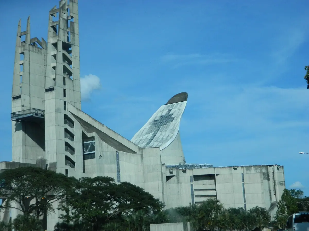 El santuario de Coromoto, de concreto gris, con su gran techo en forma de vela curvada y dos torres altas, entre árboles y un jardín