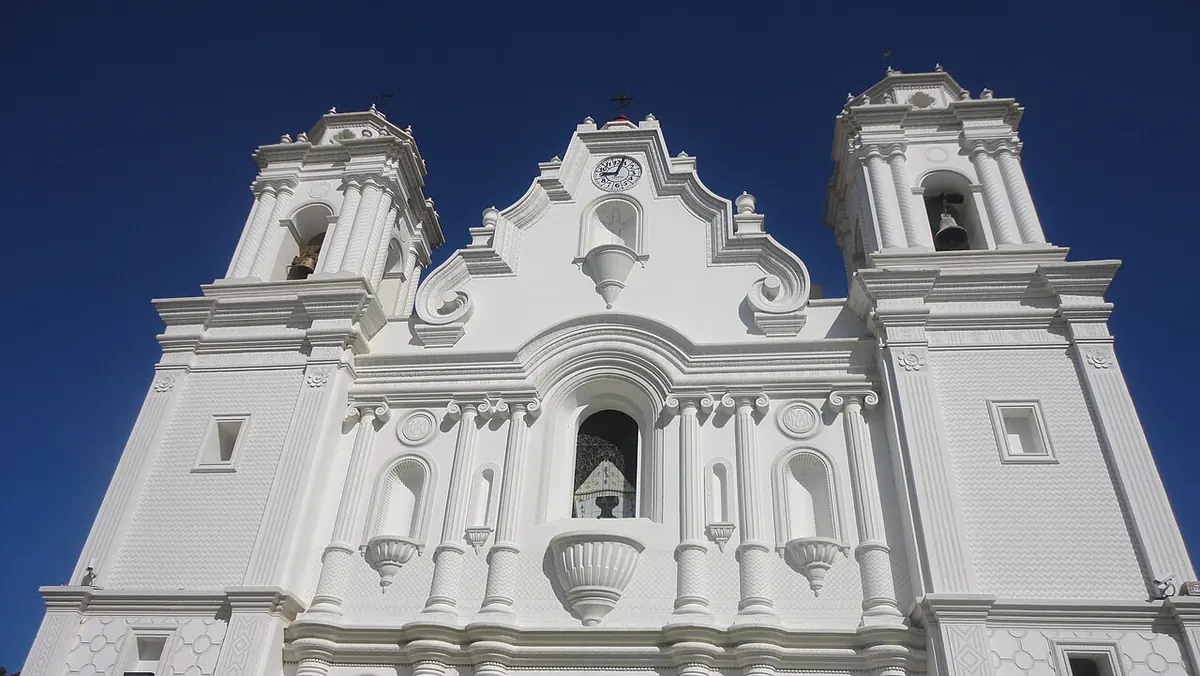 Fachada blanca del santuario de Juquila, de estilo barroco, con dos torres de campanario y un nicho central, bajo un cielo azul intenso