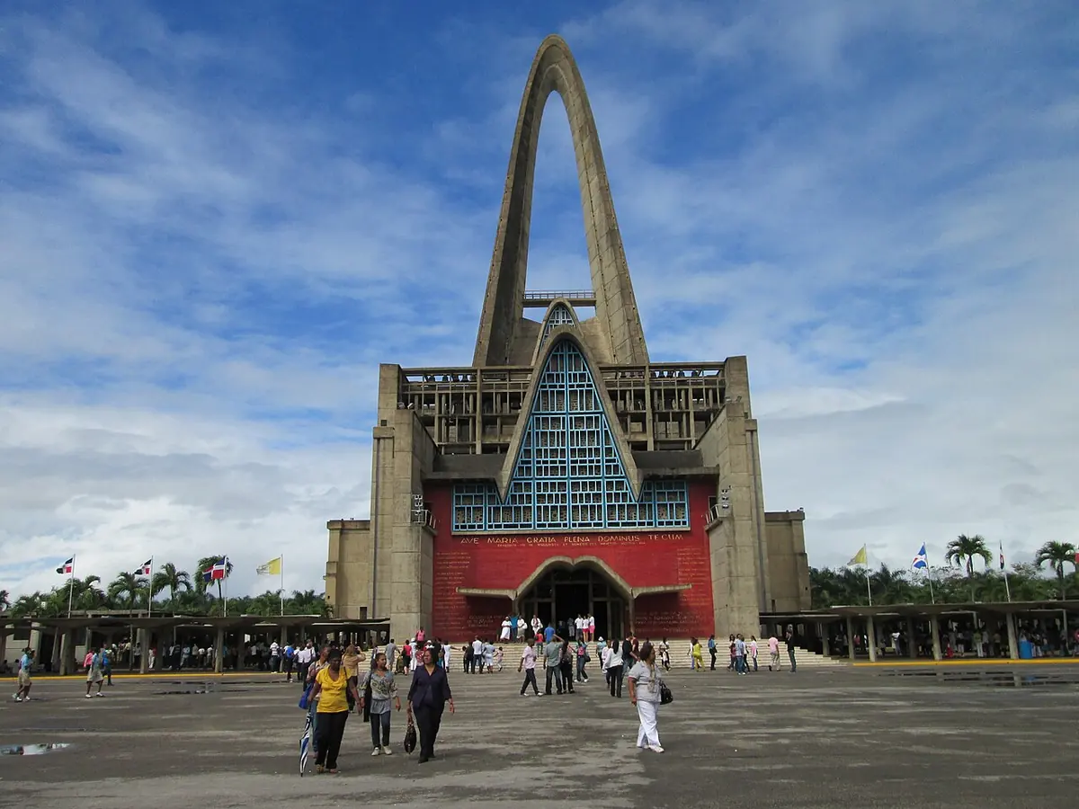 Fachada de hormigón de la basílica de Higüey, con un gran arco en punta que se eleva sobre la entrada y una vidriera azul, y peregrinos en la explanada