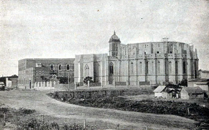 Fotografía antigua de una gran iglesia gótica a medio construir, con el ábside levantado y sin torres, junto a un camino de tierra en el campo