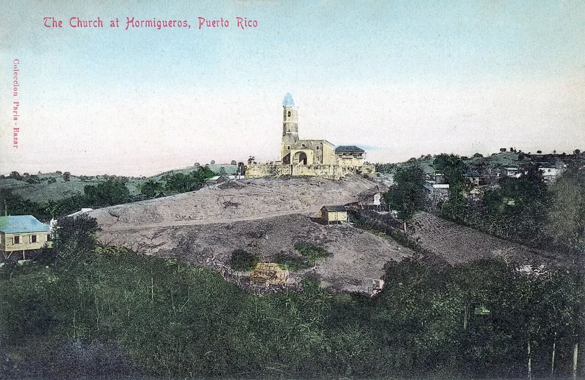 Postal antigua coloreada de la iglesia de la Monserrate de Hormigueros, con su torre, en lo alto de una loma rodeada de casas y vegetación