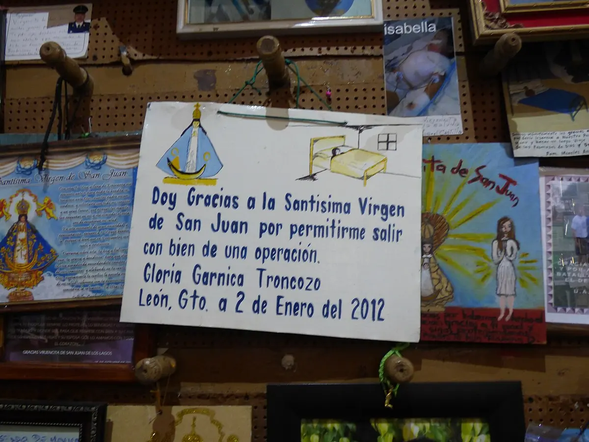 Un letrero de madera escrito a mano que da gracias a la Virgen de San Juan, con un dibujo de una cama, entre fotografías y otros exvotos colgados en la pared