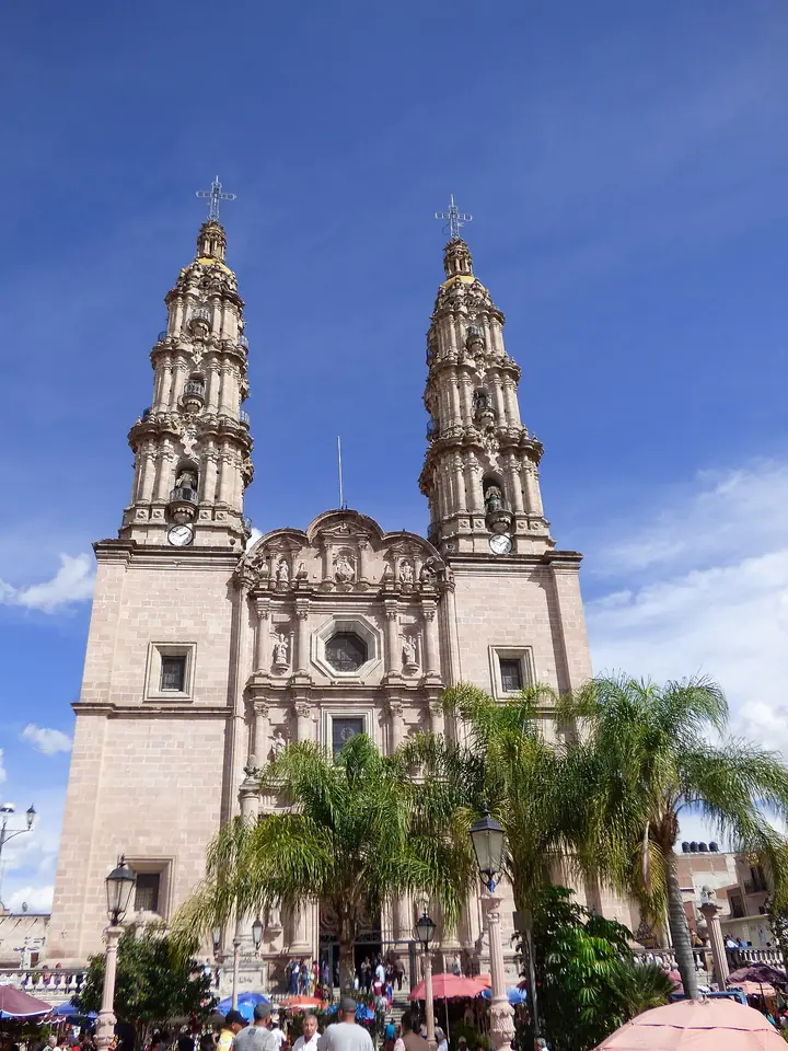 Fachada barroca de cantera rosada de la basílica de San Juan de los Lagos, con dos altas torres de campanario bajo un cielo azul y palmeras en la plaza