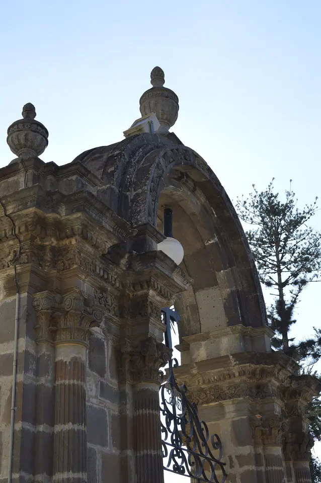 La fachada de cantera de la basílica de Talpa, con dos torres de agujas doradas y un arco de piedra labrada en el atrio, bajo un cielo azul
