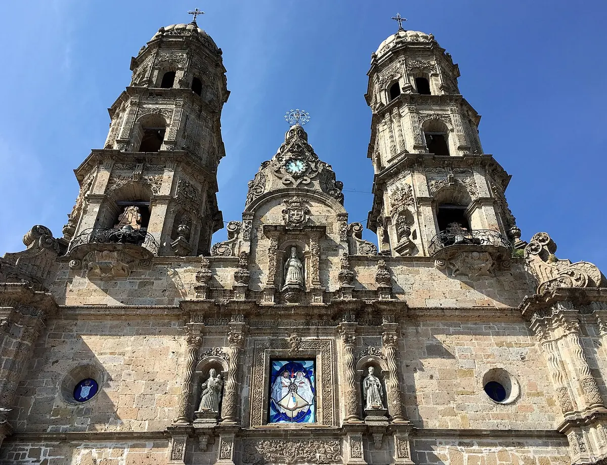 Fachada barroca de cantera de la basílica de Zapopan, con dos torres de campanario y estatuas en sus nichos, bajo un cielo azul