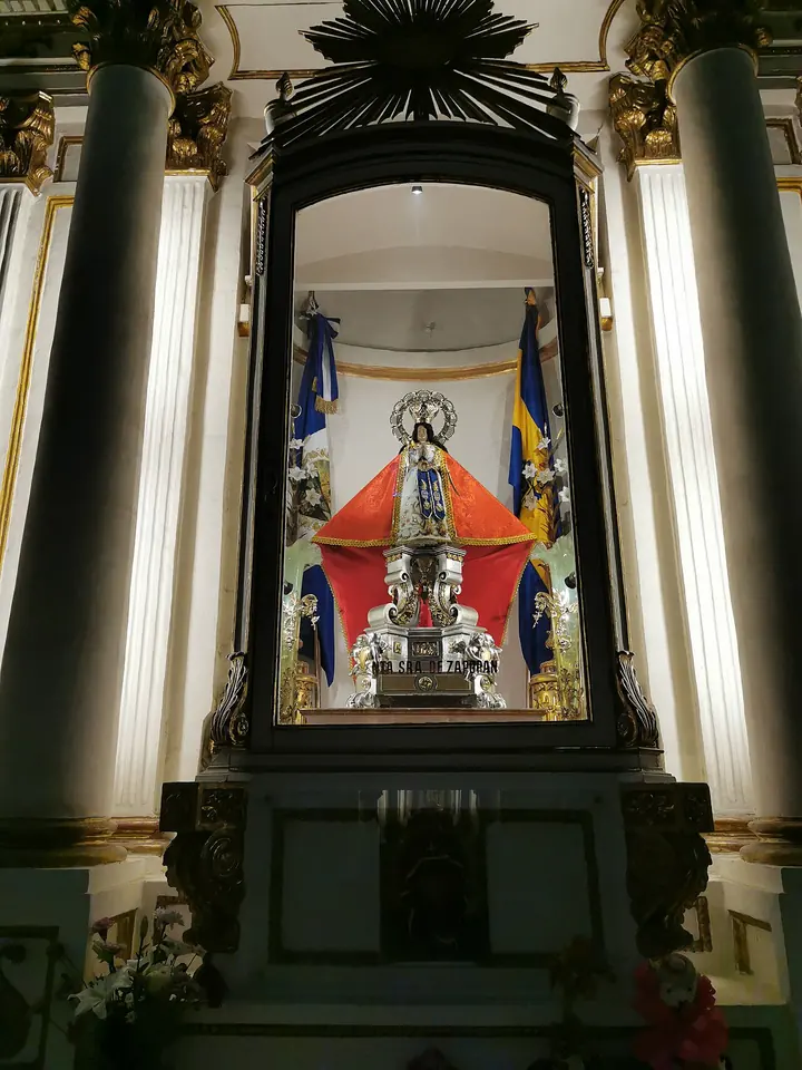 La imagen de la Virgen de Zapopan con manto rojo y corona, sobre una peana de plata dentro de un gran marco oscuro, durante su visita a la Catedral de Guadalajara