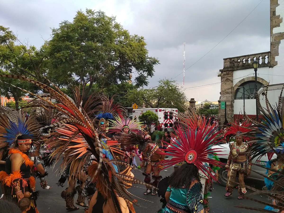 Danzantes con grandes penachos de plumas de colores bailan en la calle durante la Romería de la Virgen de Zapopan, con árboles y un edificio blanco al fondo