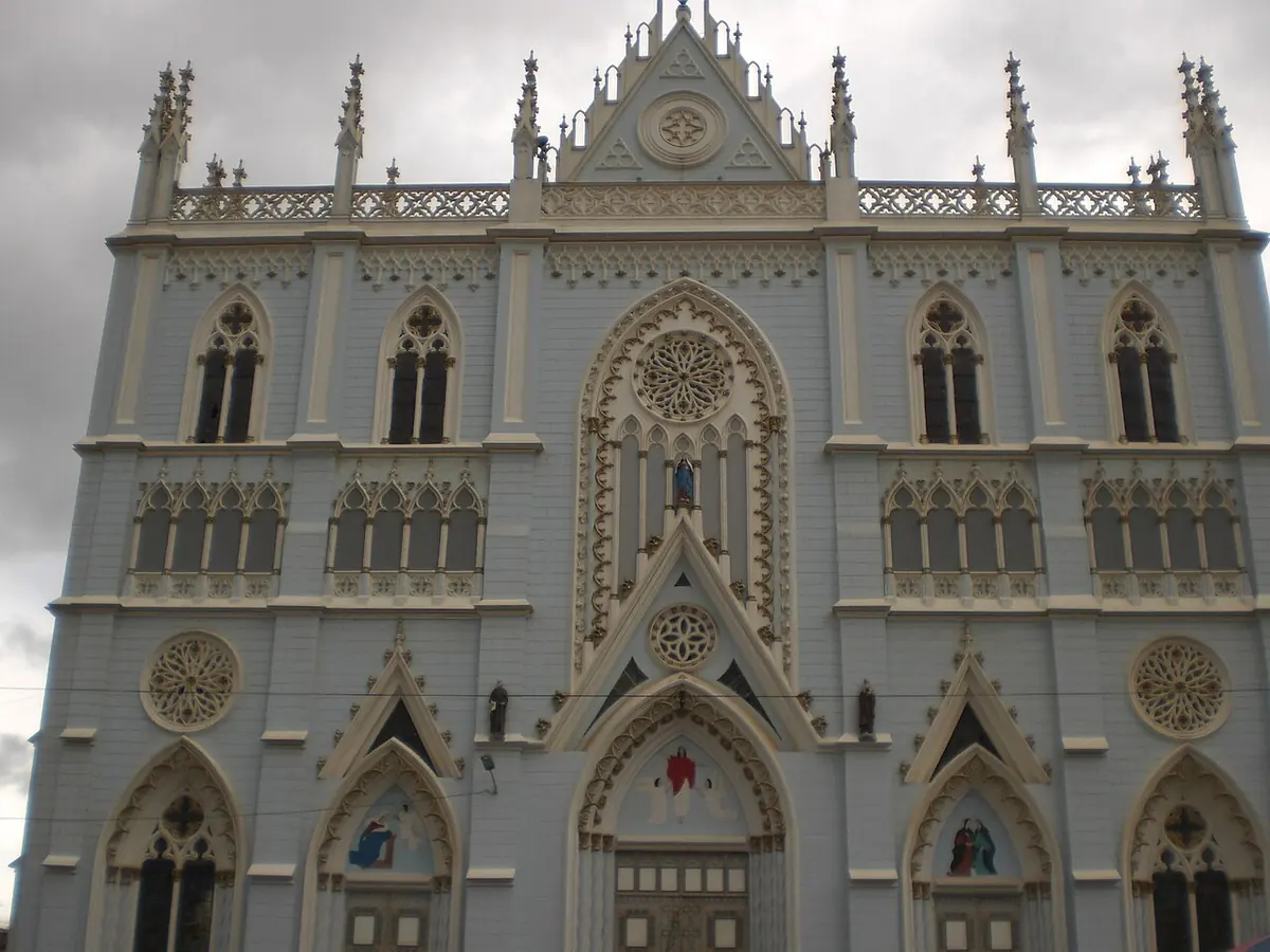 La fachada neogótica de la basílica de El Cisne, pintada de blanco y gris claro, con rosetones, ventanas ojivales y pináculos, bajo un cielo nublado